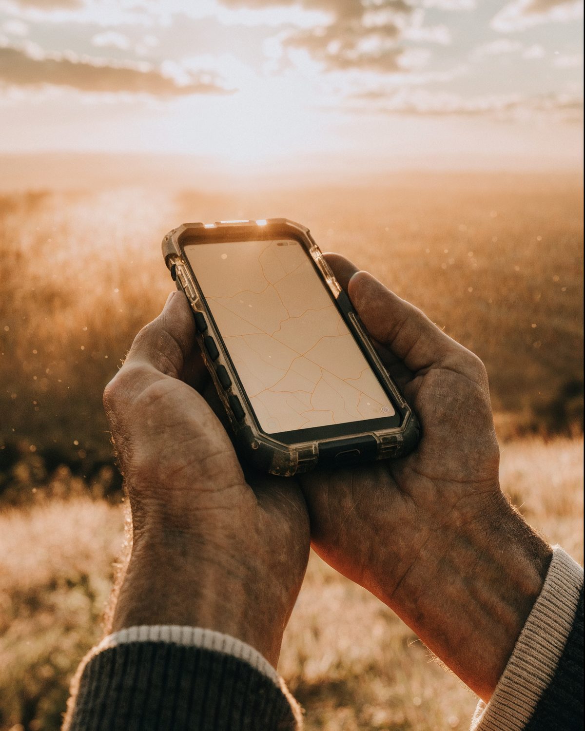 A pair of hands holding a rugged phone showing a route map, in open country at golden hour.